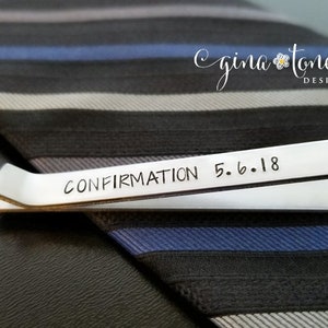 May include: Silver tie bar with the inscription "CONFIRMATION 5.6.18" on a striped tie.