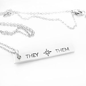 May include: Silver necklace with a bar pendant that reads "THEY THEM" with a star in between the words.