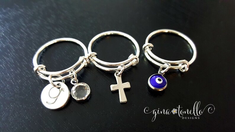 K&ouml;nnte beinhalten: Drei silberne Ringe mit Anh&auml;ngern. Der erste Ring hat einen silbernen Anh&auml;nger mit dem eingravierten Buchstaben "G", der zweite Ring hat einen silbernen Kreuzanh&auml;nger und der dritte Ring hat einen blauen Gl&uuml;cksbringer-Anh&auml;nger.