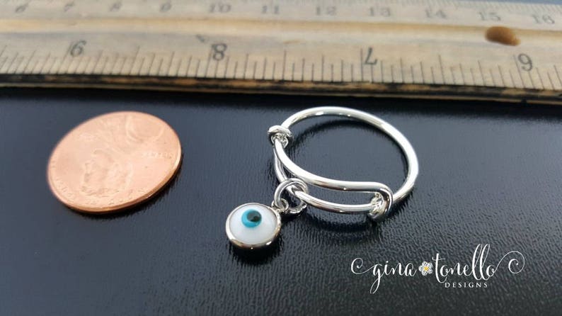 K&ouml;nnte beinhalten: Ein verstellbarer Silberring mit einem Anh&auml;nger in Form eines blauen und wei&szlig;en b&ouml;sen Auges. Der Ring liegt neben einem Centst&uuml;ck zum Gr&ouml;&szlig;envergleich.