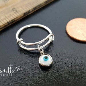 K&ouml;nnte beinhalten: Ein verstellbarer Silberring mit einem Anh&auml;nger eines wei&szlig;en b&ouml;sen Auges mit einem blauen Zentrum. Der Ring liegt neben einem US-Cent zum Gr&ouml;&szlig;envergleich.