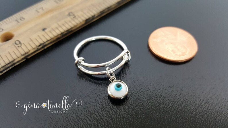 K&ouml;nnte beinhalten: Ein verstellbarer Silberring mit einem Anh&auml;nger eines wei&szlig;en b&ouml;sen Auges mit einem blauen Zentrum. Der Ring liegt neben einem US-Cent zum Gr&ouml;&szlig;envergleich.