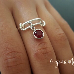 K&ouml;nnte beinhalten: Ein silberner Ring mit einem roten Edelstein, der herunterh&auml;ngt. Der Ring besteht aus miteinander verbundenen Metallteilen und hat ein schlichtes Design.