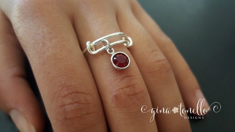 K&ouml;nnte beinhalten: Ein silberner Ring mit einem roten Edelstein, der herunterh&auml;ngt. Der Ring besteht aus miteinander verbundenen Metallteilen und hat ein schlichtes Design.
