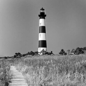 May include: Black and white photograph of a lighthouse with black and white stripes. A wooden walkway leads through tall grass towards the lighthouse. The sky is clear and the image is in black and white.