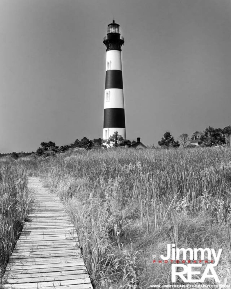 May include: Black and white photograph of a lighthouse with black and white stripes. A wooden walkway leads through tall grass towards the lighthouse. The sky is clear and the image is in black and white.