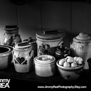 May include: Black and white photograph of a collection of antique ceramic jars and bowls. The jars vary in size and shape, some with lids and decorative patterns, including a heart design. A bowl contains several white, round objects.