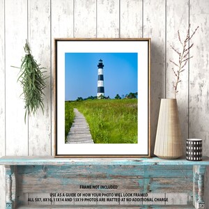 May include: Framed print of a lighthouse with black and white stripes, set against a blue sky. A wooden walkway leads through a field of green and yellow grass. The frame is brown, and the print is displayed on a distressed blue shelf.