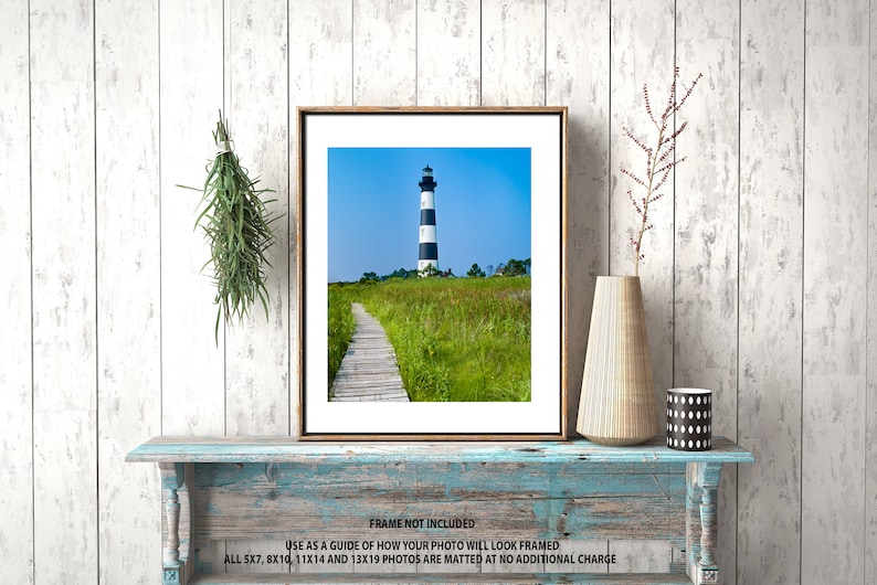May include: Framed print of a lighthouse with black and white stripes, set against a blue sky. A wooden walkway leads through a field of green and yellow grass. The frame is brown, and the print is displayed on a distressed blue shelf.