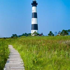 May include: A tall lighthouse with black and white stripes stands against a clear blue sky. A wooden boardwalk leads through tall green grass and reeds towards the lighthouse. The image is a scenic outdoor shot.