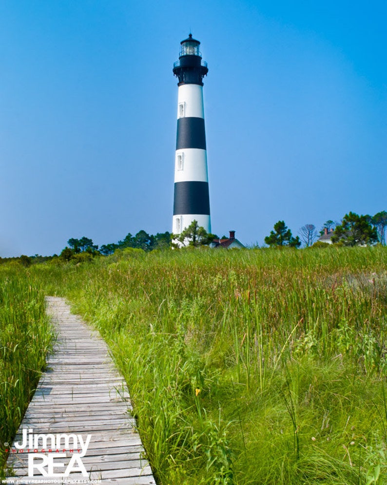 May include: A tall lighthouse with black and white stripes stands against a clear blue sky. A wooden boardwalk leads through tall green grass and reeds towards the lighthouse. The image is a scenic outdoor shot.