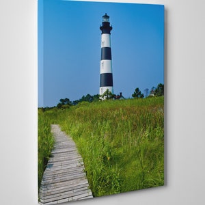 May include: A canvas print of a lighthouse with black and white stripes against a clear blue sky. A wooden boardwalk leads through tall green grass towards the lighthouse. The scene evokes a sense of tranquility and coastal charm.