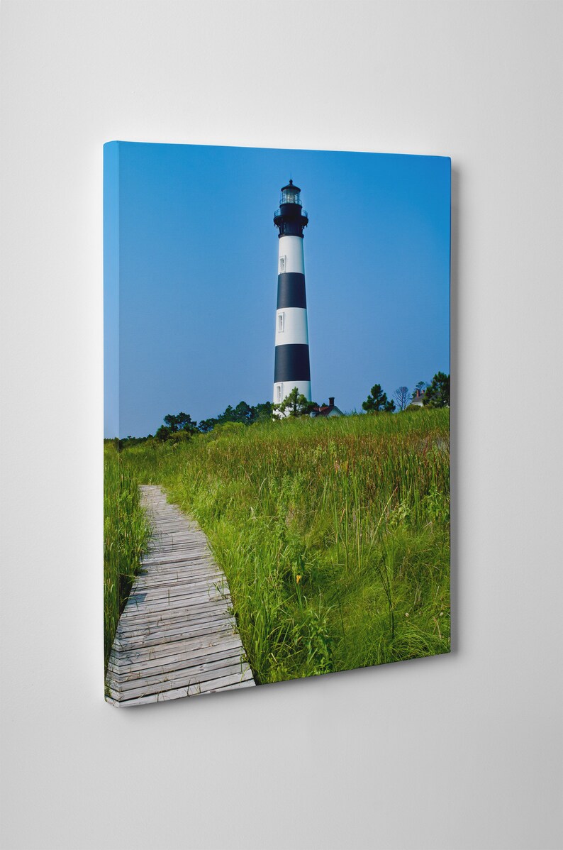 May include: A canvas print of a lighthouse with black and white stripes against a clear blue sky. A wooden boardwalk leads through tall green grass towards the lighthouse. The scene evokes a sense of tranquility and coastal charm.