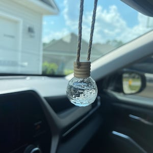 May include: A clear glass car air freshener with a faceted design hangs from a brown string. The air freshener is shaped like a sphere and has a brown wooden cap.
