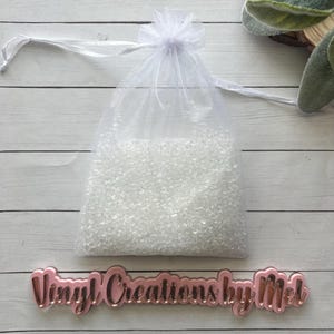 May include: A white, sheer drawstring bag filled with small, clear beads. The bag is tied with a white ribbon and sits on a white wooden surface. A pink sign with the text "Vinyl Creations by Mel" is below the bag.