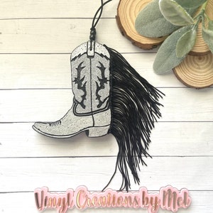 May include: A silver glitter cowboy boot with black fringe hanging from a black cord. The boot has a black design on the front. The boot is hanging from a wooden surface.