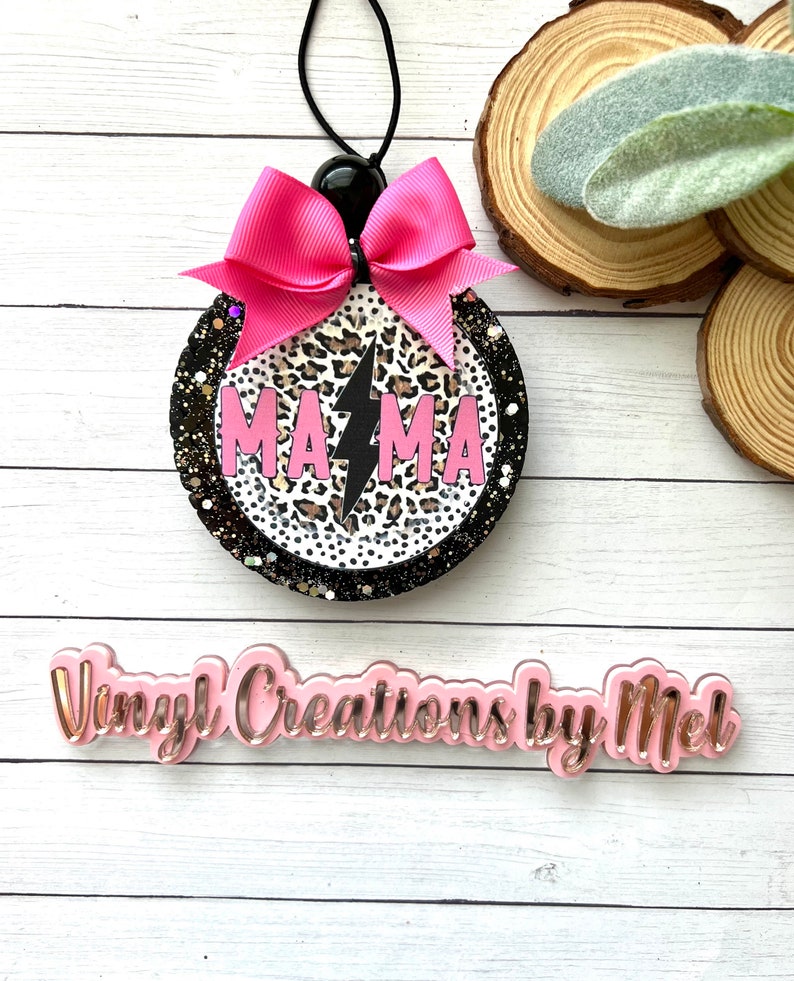 May include: A round, black and white car air freshener with a pink bow and the word "MAMA" with a lightning bolt in the center. The air freshener is made of resin and has a glitter finish.
