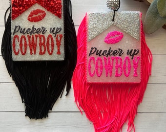 May include: Two car air fresheners with the text "Pucker Up Cowboy". One has a red glitter top and black fringe, the other has a silver glitter top and pink fringe. Each has a lip graphic.