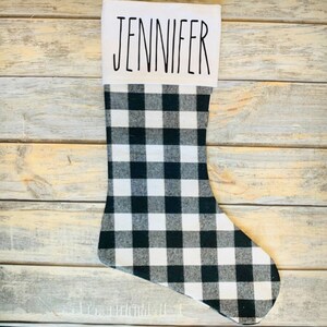 May include: A Christmas stocking with a black and white buffalo plaid pattern. The top of the stocking is white with the name "JENNIFER" printed in black. The stocking is made of fabric and is designed for the holiday season.