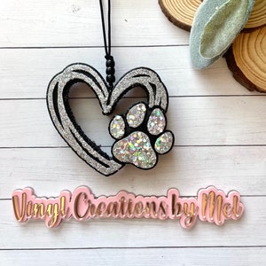 May include: A silver glitter heart-shaped ornament with a silver glitter paw print inside. The ornament is hanging from a black string. The text "Vinyl Creations by Mel" is on a pink background.