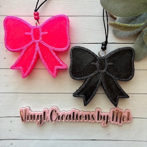 May include: Two glitter bow tie car air fresheners, one pink and one black, hanging from black cords with black beads. The text "Vinyl Creations by Mel" is visible below the bows.