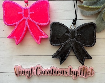 Könnte beinhalten: Zwei glitzernde Schleifen-Auto-Lufterfrischer, einer pink und einer schwarz, hängen an schwarzen Schnüren mit schwarzen Perlen. Der Text "Vinyl Creations by Mel" ist unter den Schleifen sichtbar.