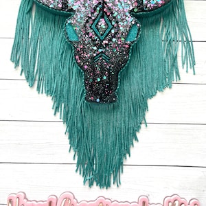 May include: A decorative wall hanging featuring a bull skull design. The skull is black with glitter accents and turquoise details. Turquoise fringe hangs below the skull. A small, sparkly ball hangs above the skull.