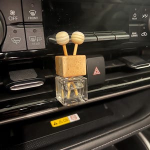 May include: A car air freshener with a clear glass cube base and a wooden block top, topped with two light-colored wooden spheres. The freshener is placed on a car dashboard, near the air vents.