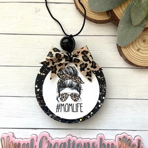 May include: A round car air freshener with a black string and a black bead. The air freshener has a white center with a woman wearing sunglasses and a messy bun, with the text "#MOMLIFE". A leopard print bow is attached.