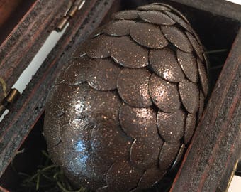 May include: A dark brown dragon egg with a scaly texture, resting in a wooden box lined with green moss.