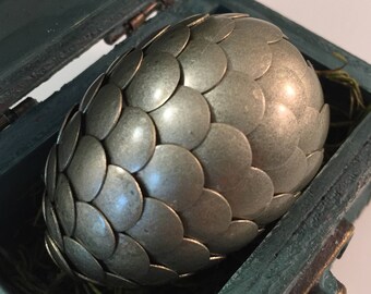 May include: A metallic silver egg with a scale-like pattern rests inside a teal wooden box lined with green moss. The egg is oval-shaped and has a textured surface, creating a unique decorative piece.