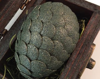 May include: A green dragon egg with a scaly texture is nestled in a wooden box lined with green moss.