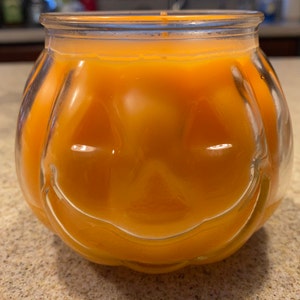 Pode incluir: Um porta-velas de vidro transparente em forma de ab&oacute;bora com uma vela laranja brilhante dentro. A vela tem um rosto esculpido com um sorriso.