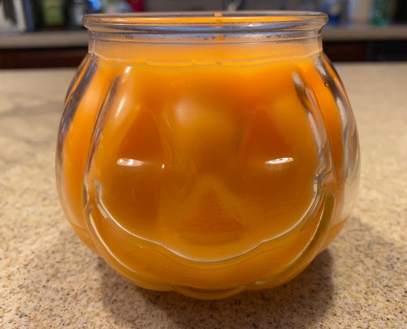 Pode incluir: Um porta-velas de vidro transparente em forma de ab&oacute;bora com uma vela laranja brilhante dentro. A vela tem um rosto esculpido com um sorriso.