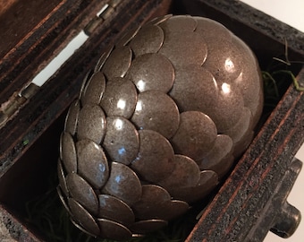 May include: A silver dragon egg with a scaly texture sits inside a wooden box with a green moss lining.