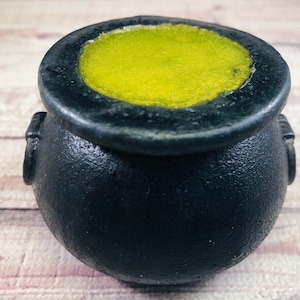 May include: A black cauldron with a textured surface, filled with a bright yellow-green liquid. The cauldron has two small handles on the sides and a slightly rough rim. The image is taken from a close-up angle.