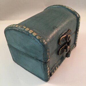 May include: A small teal blue wooden box with a gold trim and a brass latch. The box is closed and sitting on a white surface.