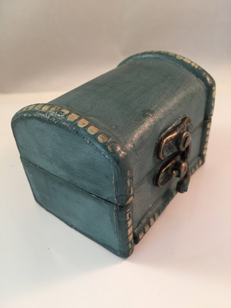 May include: A small teal blue wooden box with a gold trim and a brass latch. The box is closed and sitting on a white surface.