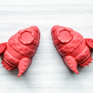 May include: Two bright red rocket-shaped silicone molds. The molds have a detailed design with a round window, a grid pattern, and stars.