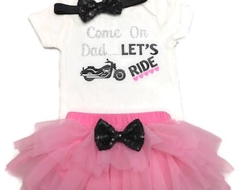 Könnte beinhalten: Ein weißer Baby-Strampler mit dem Text "Come On Dad... LET'S RIDE" und einer schwarzen Schleife. Der Strampler wird mit einem rosa Tüll-Tutu mit einer schwarzen Schleife kombiniert.