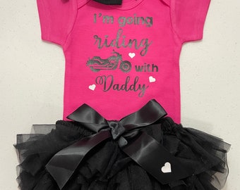Könnte beinhalten: Rosa Baby-Strampler mit dem Text "I'm going riding with Daddy" und einer Motorradgrafik. Der Strampler wird mit einem schwarzen Tüll-Tutu mit einer schwarzen Satin-Schleife kombiniert.