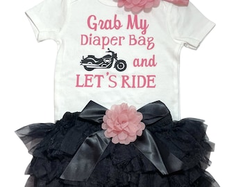 May include: A white baby outfit with a pink floral headband. The outfit includes a white onesie with the text "Grab My Diaper Bag and LET'S RIDE" and a black tutu skirt with a pink flower and bow.