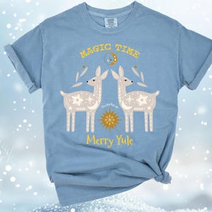 May include: A light blue t-shirt with a graphic of two stylized deer with a yellow star and the text "Magic Time" and "Merry Yule".