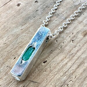 May include: A silver pendant necklace featuring a rectangular pendant with a vibrant green gemstone. The pendant is suspended from a delicate silver chain. The necklace is displayed on a wooden surface.