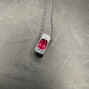 May include: A silver necklace with a rectangular pendant, showcasing a vivid, oval-cut red gemstone. The pendant hangs from a fine chain. The jewellery is set against a dark background.