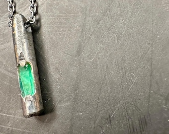 May include: A silver-toned pendant necklace featuring a rectangular, hollowed-out design. A vibrant green gemstone is visible within the pendant. The necklace has a dark chain, creating a striking contrast. The jewelry is displayed against a dark background.