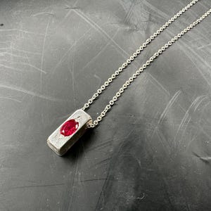 Puede incluir: Un collar de plata con un colgante rectangular con una gema roja vibrante. El colgante tiene una textura y la cadena es delicada. La joya se muestra sobre un fondo oscuro.