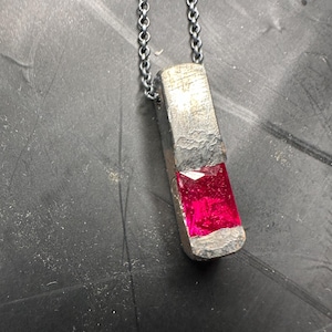 Op de afbeelding: Een zilverkleurige hangerketting met een rechthoekige, getextureerde hanger met een levendige, rechthoekige, robijnrode edelsteen. De hanger hangt aan een delicate, zilveren ketting, wat een opvallend contrast creëert. Het sieraad wordt tegen een donkere achtergrond getoond.