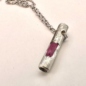 Op de afbeelding: Zilverkleurige ketting met een cilindervormige hanger. De hanger heeft een textuur en een rechthoekige inleg van roze edelsteen. De hanger hangt aan een fijne ketting.