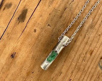 May include: A silver-toned pendant necklace featuring a cylindrical pendant with a section of vibrant green stones. The pendant hangs from a delicate chain. The necklace is displayed on a wooden surface.